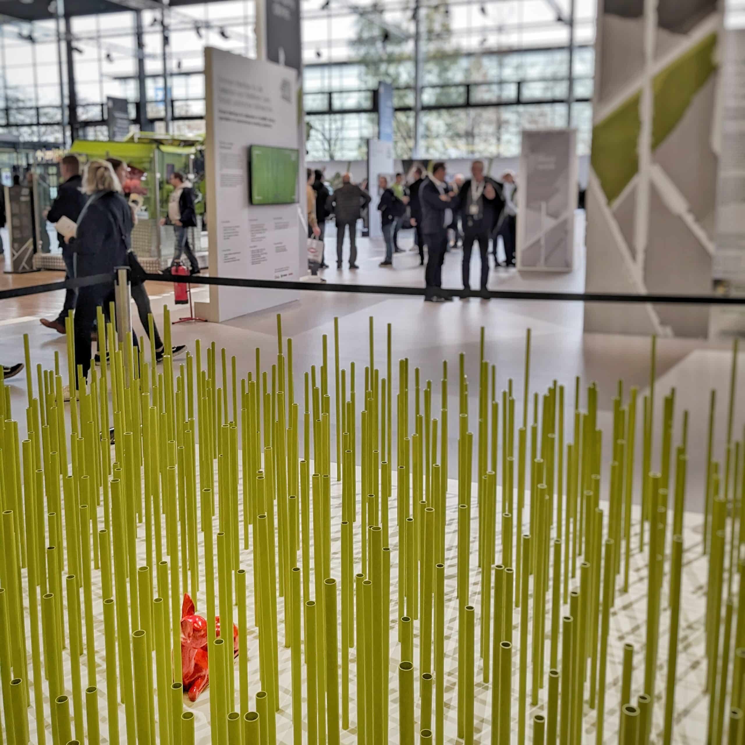 Bild einer Installation künstlicher Getreide-Halme mit einer Figur eines Rehkitzes darin. Im Hintergrund ist eine Halle der Agritechnica 2023 zu erkennen, in der mehrere Personen an verschiedenen Ständen stehen.