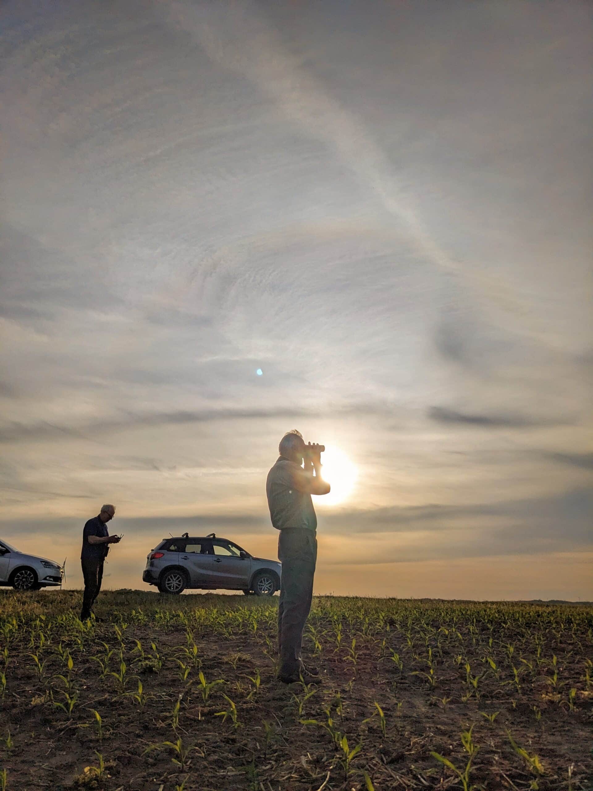 Bild von 2 Personen, die auf einem Feld stehen. Die eine Person schaut durch ein Fernglas, während die andere Person im Hintergrund auf einen Drohnencontroller guckt. Hinter den beiden stehen zwei Autos am Feldrand.