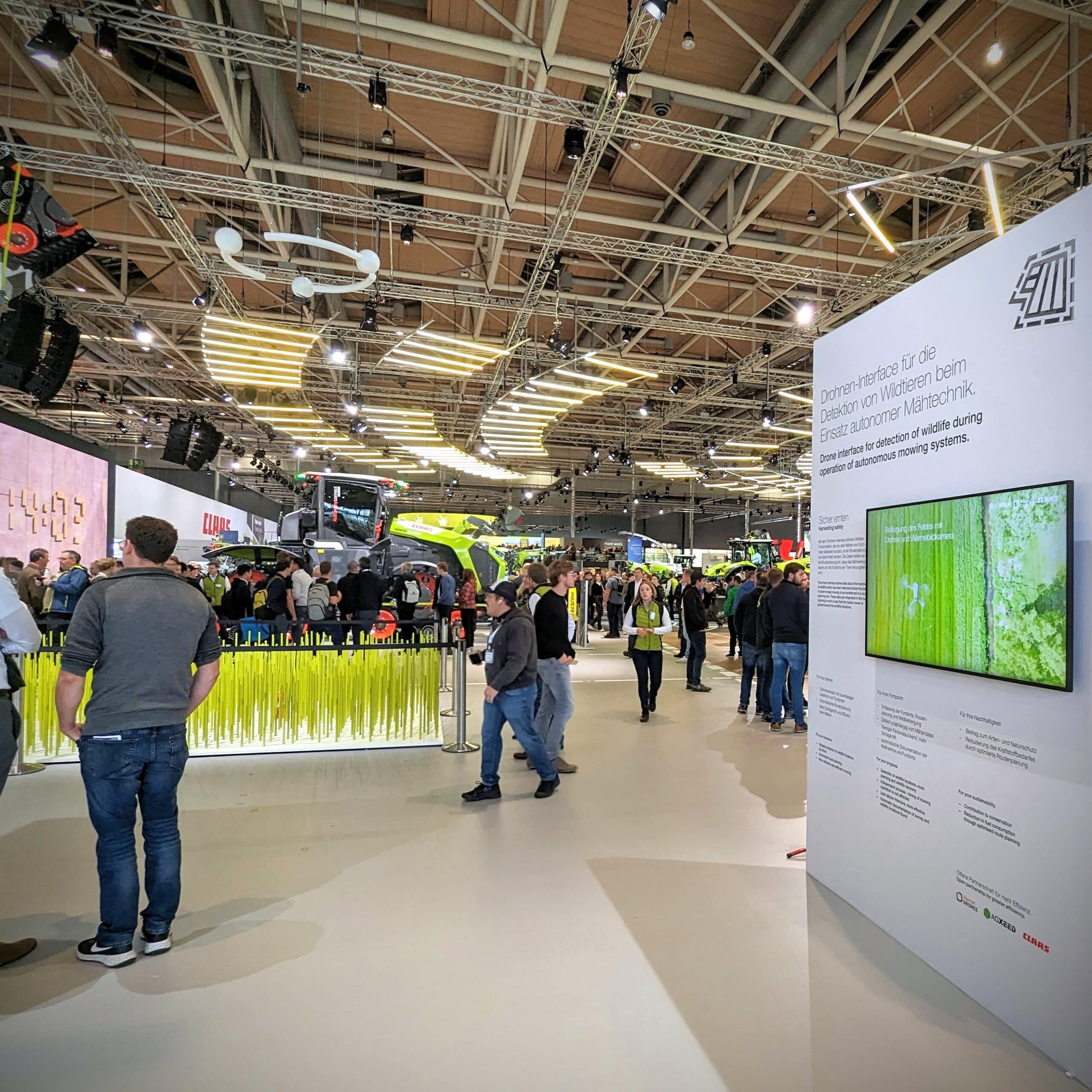 Messestand von CLAAS auf der Agritechnica 2023, mit einem abstraktem Feld mit einem Rehkitz auf der linken Seite und einer Infowand auf der rechten Seite. Ein großer Traktor ist im Hintergrund zu sehen. Die Messe ist gut besucht.