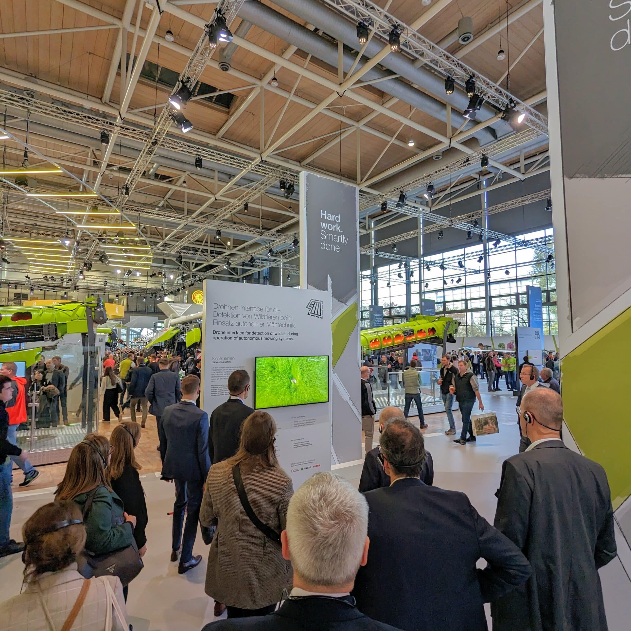 Menschen stehen um einen Messestand auf der Agritechnica 2023, welcher ein Drohnen-Interface für die Detektion von Wildtieren beim Einsatz autonomer Mähtechniken zeigt.