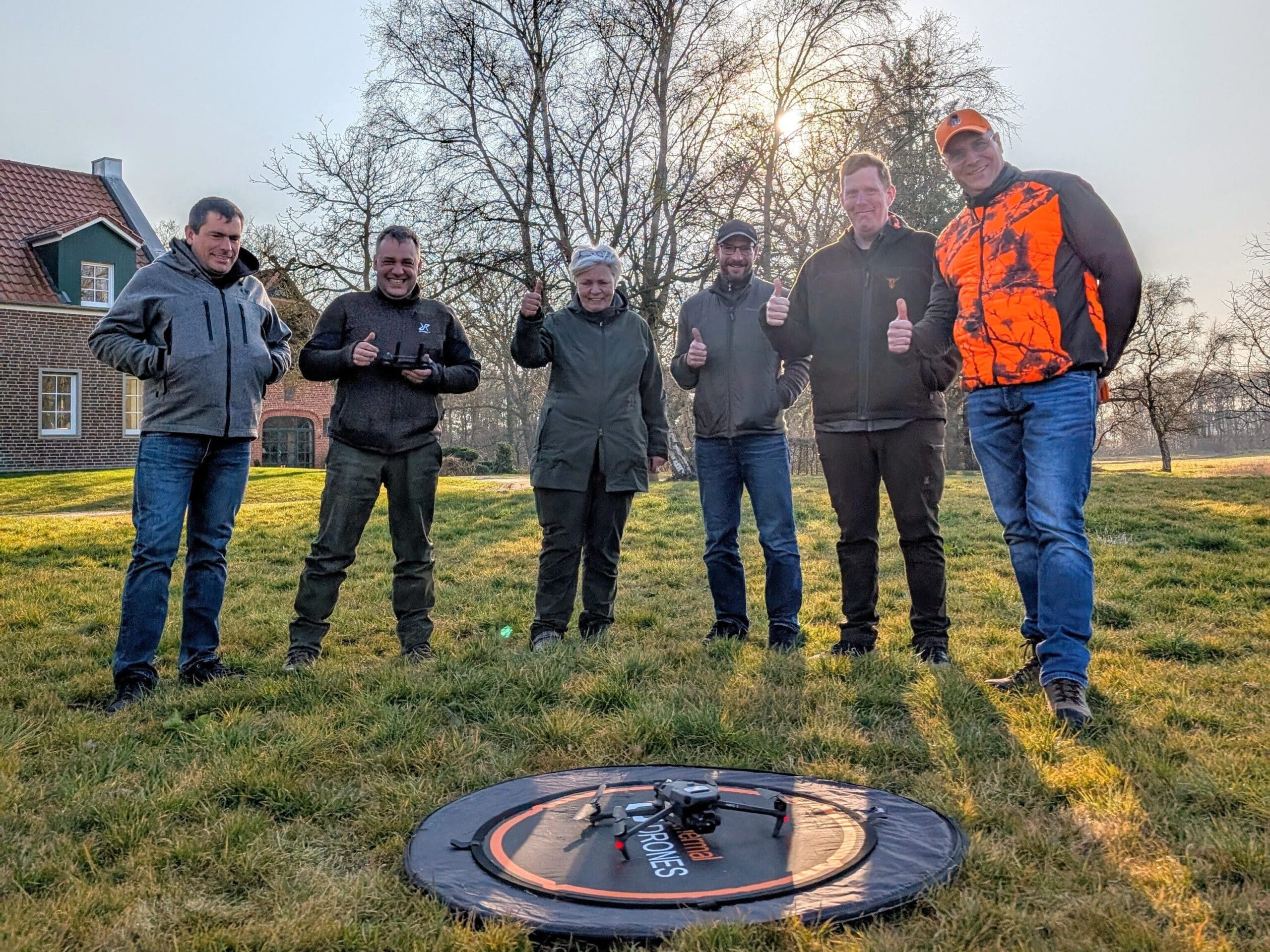 Eine Gruppe von Menschen, die auf einer Wiese steht. Es sind 6 Personen, die im Bogen um eine Wärmebilddrohne stehen, die am Boden auf einem thermal DRONES Landepad steht. Die Personen lächeln, zeigen den rechten Daumen nach oben und schauen fröhlich in die Kamera. Im Hintergrund sind einige Bäume und ein Haus zu sehen.