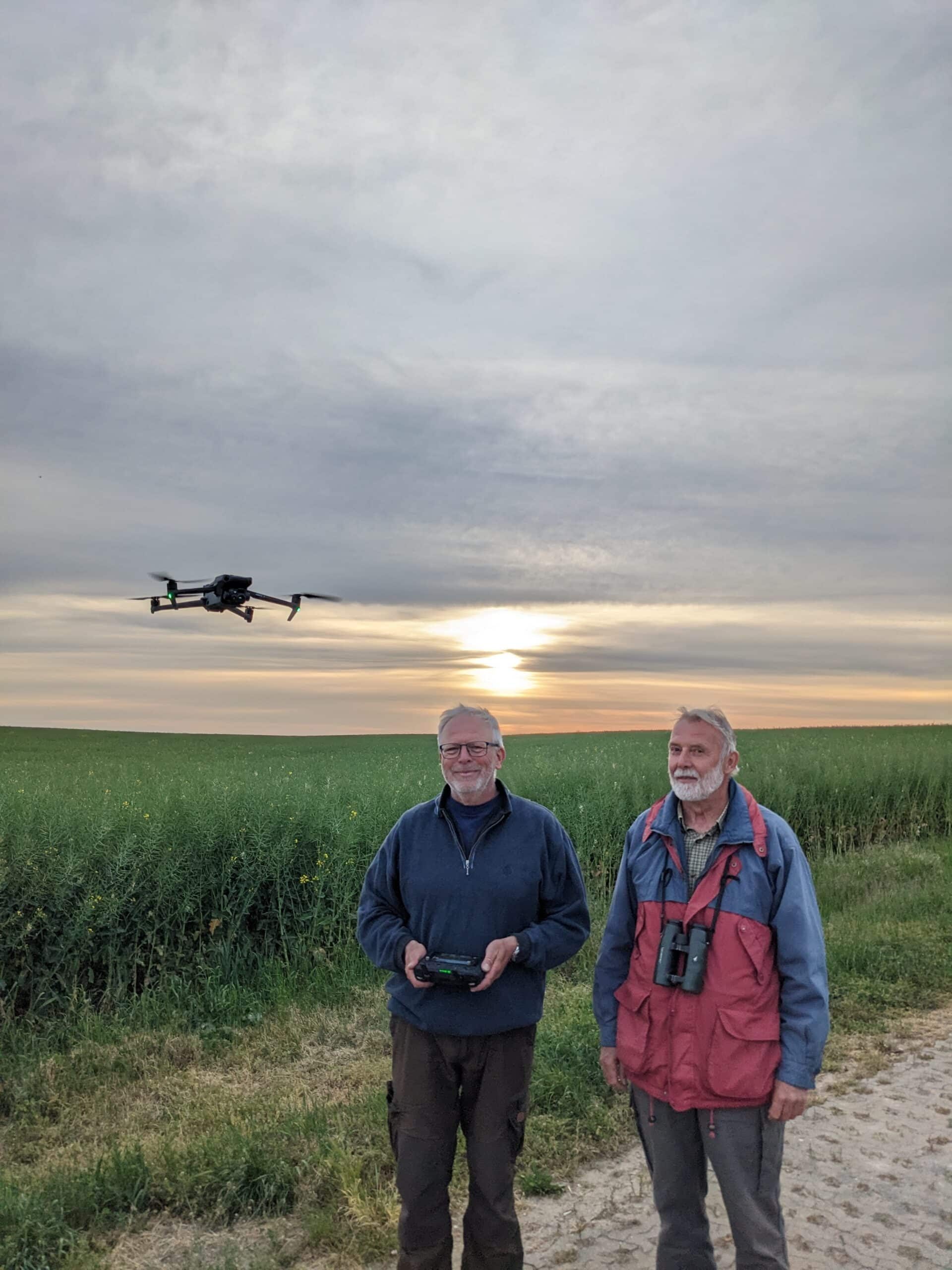 Bild von zwei Personen, die vor einem Feld stehen. Links neben den beiden fliegt in geringer Höhe eine Drohne. Die Person auf der linken Seite hält den dazugehörigen Drohnencontroller in der Hand. Die Person auf der rechten Seite steht daneben und sieht in die Kamera. Sie trägt ein Fernglas um den Hals. Im Hintergrund sieht man die Sonne, die hinter einer Wolkendecke verborgen ist.