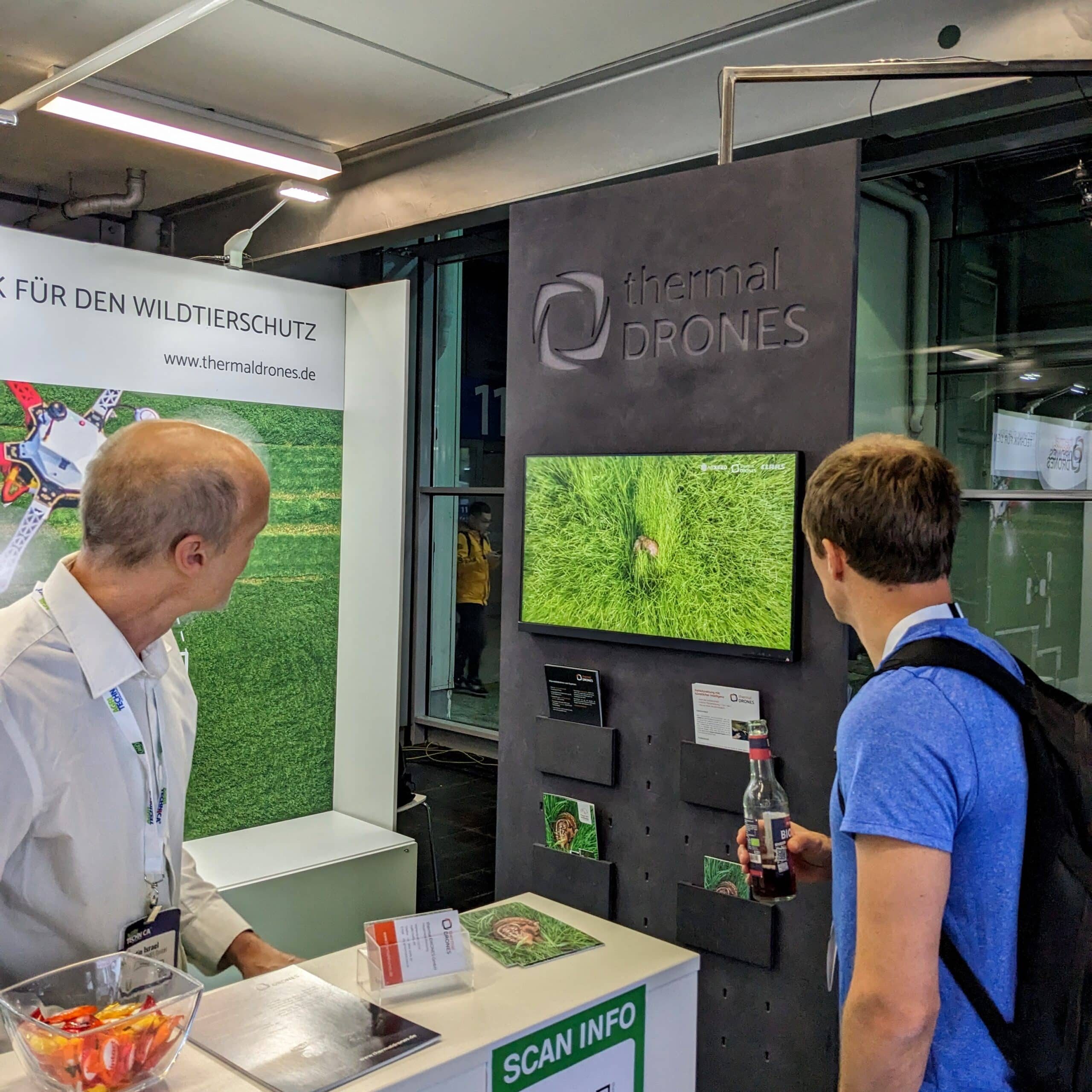 Bild vom thermal DRONES-Messestand auf der Agritechnica 2023. 2 Personen stehen an einem Tisch und schauen gemeinsam auf einen Bildschirm, auf dem eine Drohnenaufnahme von einem Rehkitz im Feld zu sehen ist.