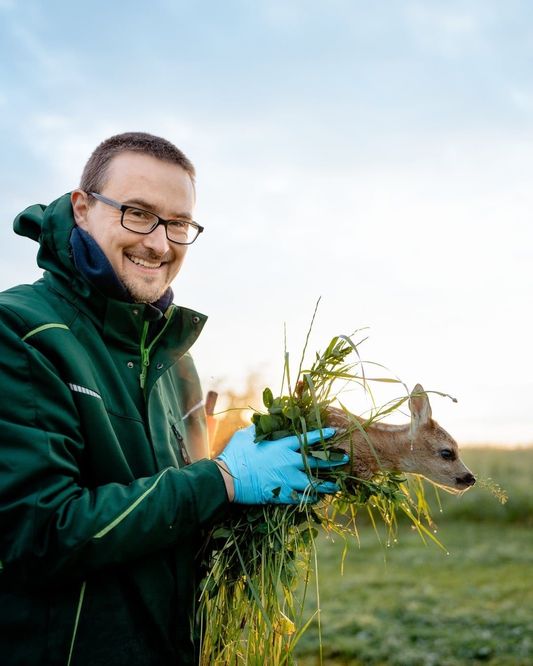 Ein Bild von einem Mann, der draußen auf einer Wiese steht. Er hält ein Rehkitz mit Gras in seinen behandschuhten Händen und lächelt in die Kamera. Im unscharfen Hintergrund sieht man ein gemähtes Feld und den blauen Himmel sowie die Strahlen der aufgehenden Sonne.