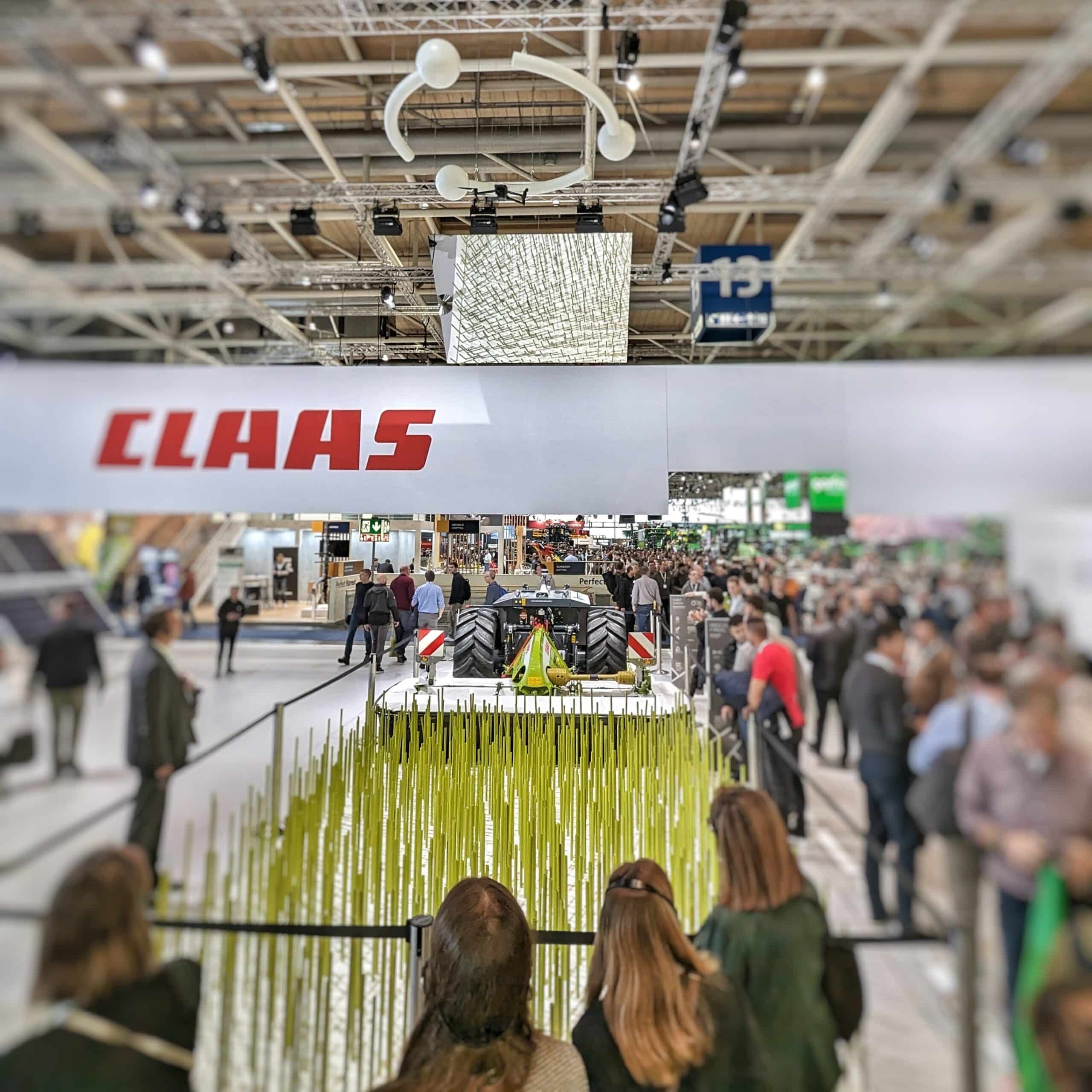 Bild eines AgXeed-Mähroboters hinter einer Installation eines künstlichen Feldes innerhalb einer Messehalle der Agritechnica 2023. Um die Installation befinden sich mehrere Personen. Im Hintergrund ist die restliche Messehalle zu erkennen.