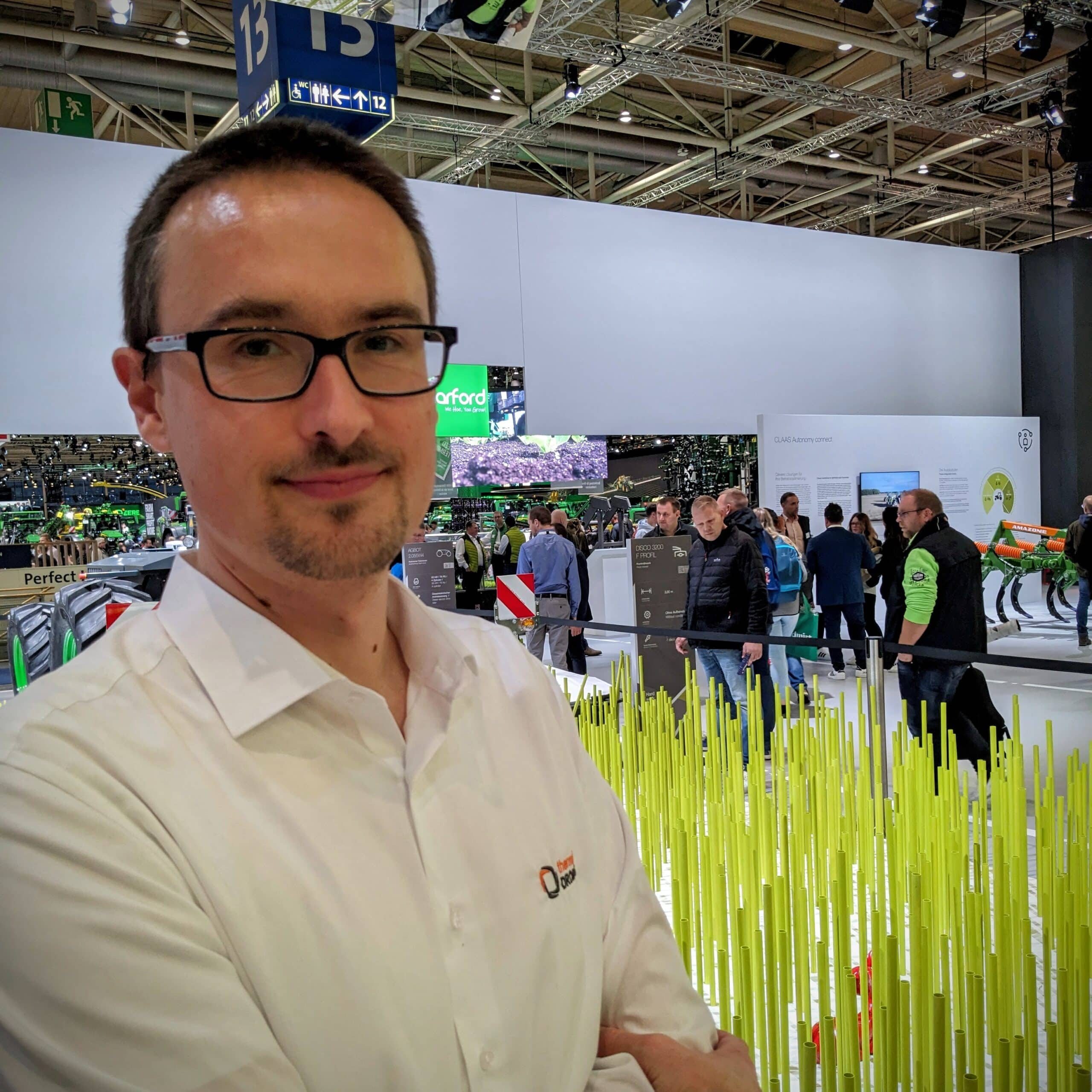 Selfie von Tobias Dahms auf der Agritechnica 2023. Im Hintergrund sind mehrere Personen an Messeständen zu sehen.