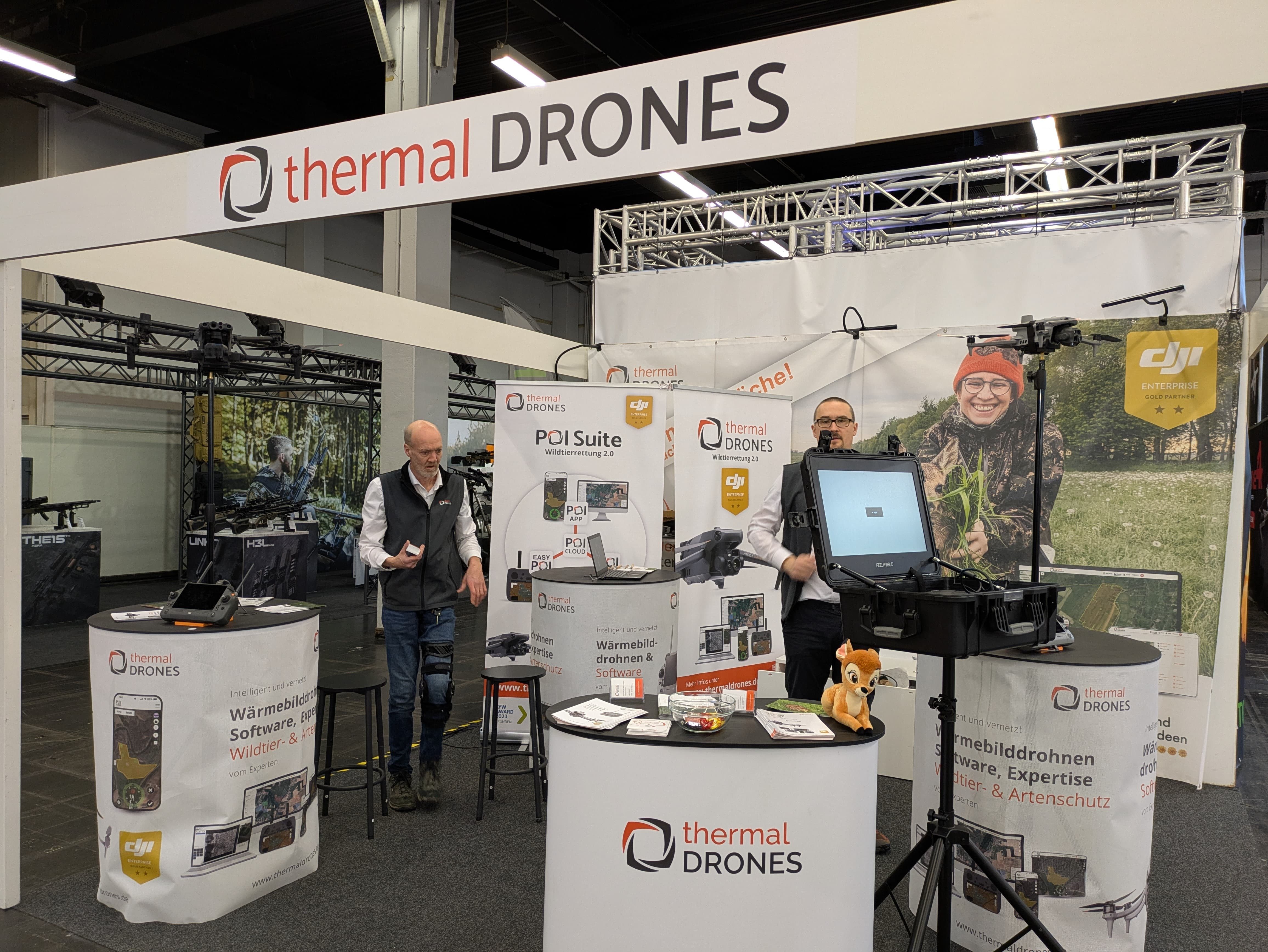 Aufnahme eines Messestandes von thermal DRONES auf der Jagd & Hund in Dortmund. Der Stand beinhaltet mehrere Tische, Plakate, Rollups und Aufsteller mit unterschiedlichen Abbildungen rund um Rehkitzrettung und die POICLoud. Auf einem Stativ steht ein Monitorkoffer, auf den das Bild der Drohne übertragen wird. Darüber hinaus sind mehrere Drohnen auf Stativen aufgestellt. Hinter den Tischen stehen 2 männliche Personen aus dem thermal DRONES - Team