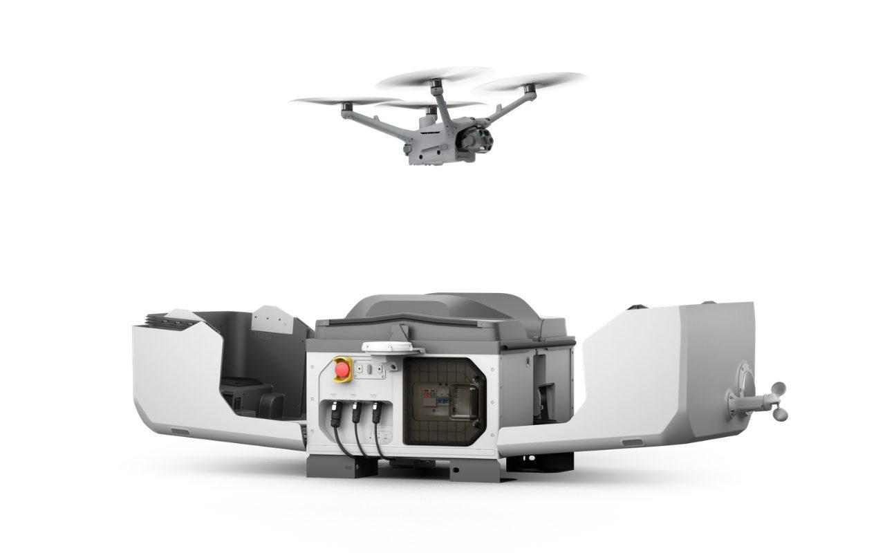 Eine DJI Matrice 4TD schwebt über der geöffneten DJI Dock 3, freigestellt auf weißem Grund. Die Abdeckung ist aufgeklappt, vorn sitzt ein roter Not-Aus-Knopf.