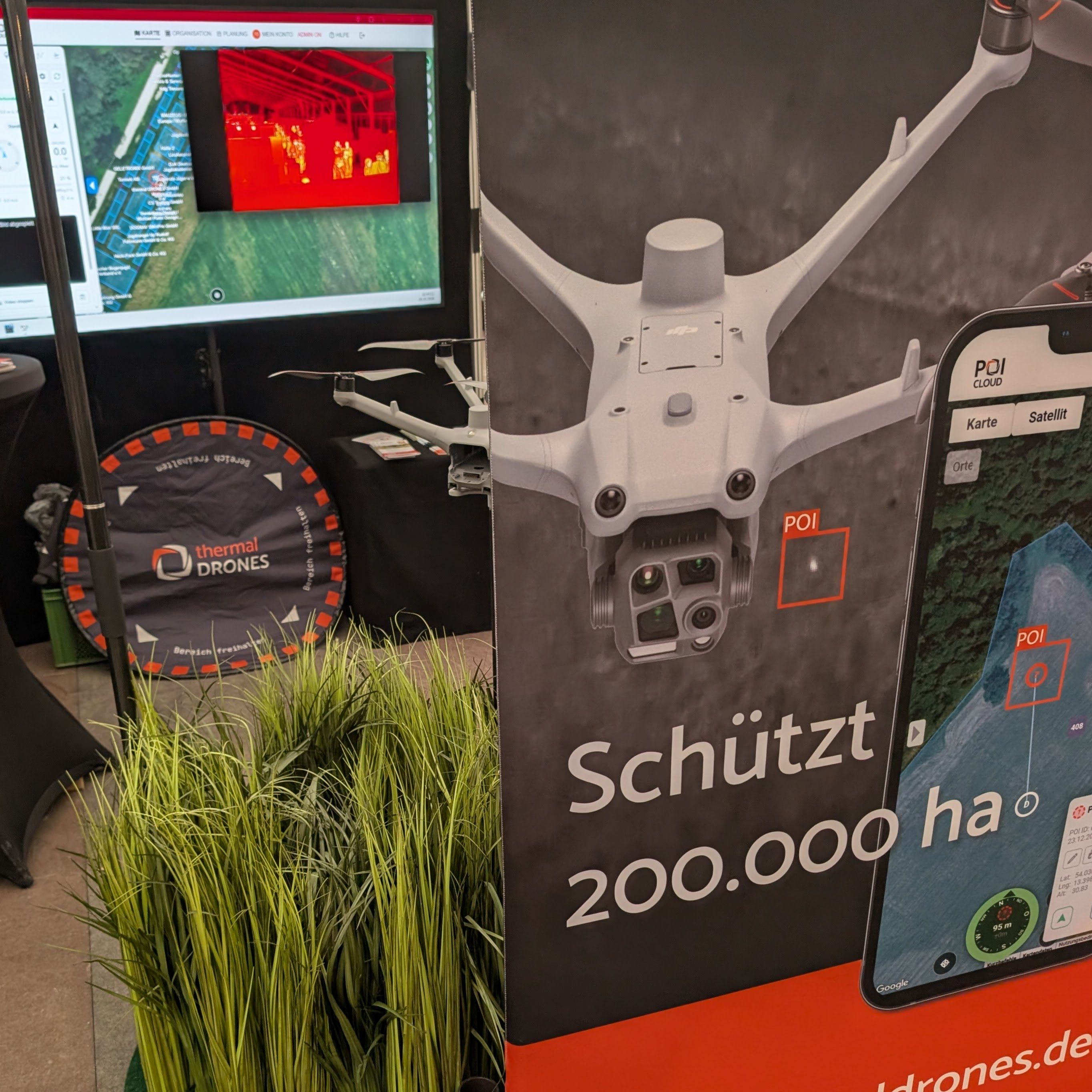 Messestand von thermal DRONES: Roll-up mit Drohne, POICloud auf dem Smartphone und dem Slogan „Schützt 200.000 ha“; dahinter ein Monitor mit Karte und Wärmebild, ein Landepad und Kunstgras